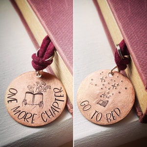 May include: Two copper-colored round bookmarks with black etched designs. One says "ONE MORE CHAPTER" with a book and flowers. The other says "GO TO BED" with a book and stars. Both have a burgundy ribbon attached.
