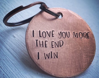 May include: A copper-colored, round keychain tag with the engraved text "I LOVE YOU MORE THE END I WIN". The tag is attached to a bronze-colored split ring. The background is a light blue and white surface.