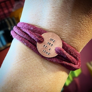 Può includere: Un braccialetto in tessuto bordeaux con un ciondolo circolare color rame. Il ciondolo è stampato con le parole "ONE DAY AT A TIME" in un semplice carattere. Il braccialetto è avvolto attorno a un polso.
