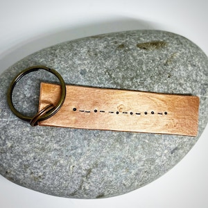 Può includere: Un portachiavi rettangolare color rame con un portachiavi color bronzo. Il portachiavi presenta una serie di punti e trattini, che rappresentano il codice Morse. Il portachiavi è appoggiato su una pietra grigia.