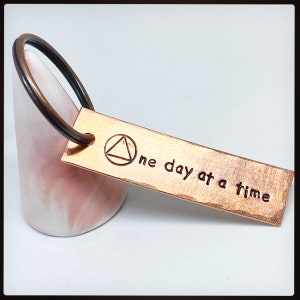 Può includere: Un portachiavi in metallo color rame con una texture martellata. Il portachiavi ha un anello circolare e un'etichetta rettangolare con il testo "one day at a time" e un simbolo di triangolo in un cerchio.