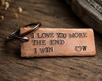 May include: A rectangular copper keychain with a hammered texture and a dark metal ring. The surface is stamped with the black text: I LOVE YOU MORE, THE END, I WIN, followed by a heart symbol and the letter W. The metal has a warm, reddish-brown tone.