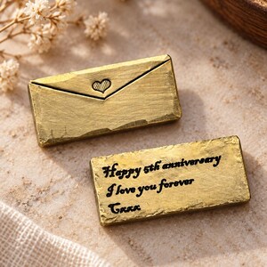 May include: Two rectangular gold-toned metal pieces with a textured finish. One piece features an engraved envelope design with a small heart. The other piece is engraved with the text: Happy 5th anniversary, I love you forever, Gxxx.