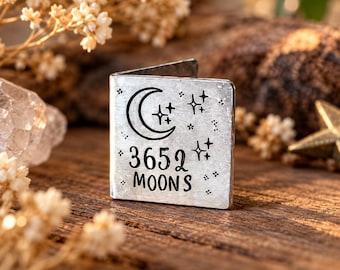 Op de afbeelding: Een vierkante boekenlegger van zilverkleurig metaal met een gevouwen rand. Het vooroppervlak is voorzien van een gegraveerde halve maan, verschillende vierpuntige sterren en de tekst 3652 MOONS in een zwart, schreefloos lettertype.
