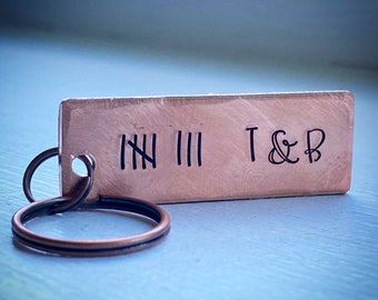 May include: A copper rectangular keychain with black markings. The keychain has tally marks and the letters "T & B" stamped on it. The keychain is attached to two metal rings.