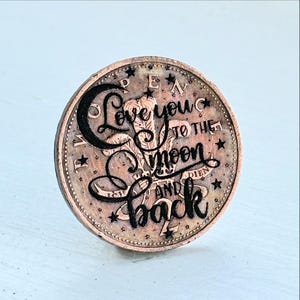 May include: A copper-colored coin with the words "Love you to the moon and back" in a decorative script. The coin has a detailed edge and small star accents around the text.