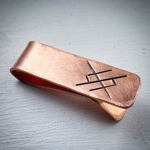 May include: A copper money clip with a brushed finish. The clip has a geometric design cut into the metal. The clip is curved and has a simple, functional design.