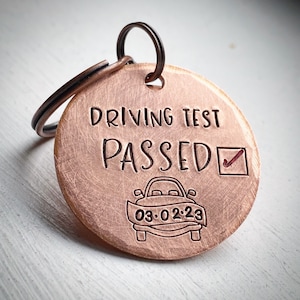 May include: A copper-colored round keychain with the words "DRIVING TEST PASSED" engraved on it. A car graphic and the date "03.02.23" are also engraved. The keychain has a dark brown key ring attached.