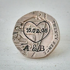 May include: A silver twenty pence coin with a heart-shaped engraving. Inside the heart is the date 15.02.05 and the initials A & B. The coin has a textured surface and the words "TWENTY PENCE" are visible.