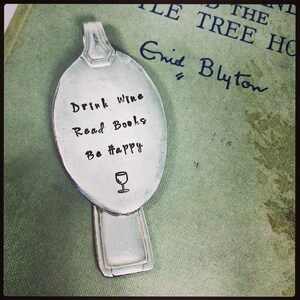 May include: A silver spoon bookmark with the words "Drink Wine Read Books Be Happy" stamped on it, and a wine glass illustration. The bookmark rests on a green book titled "The Little Tree House" by Enid Blyton.