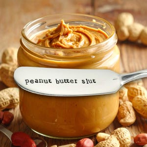 Puede incluir: Un tarro de mantequilla de cacahuete con un cuchillo de acero inoxidable que dice "peanut butter slut". El tarro está rodeado de cacahuetes y mitades de cacahuetes. La mantequilla de cacahuete es de color marrón claro.