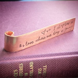 Könnte beinhalten: Ein kupferfarbener Lesezeichen mit gebogenem Ende liegt auf einem lila Buch. Der Lesezeichen hat den eingravierten Text "7 Kapitel und ich liebe, wo unsere Geschichte hingeht" mit Herzsymbolen.