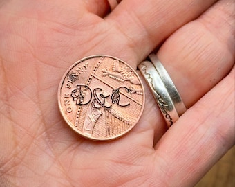 May include: A copper penny featuring the engraved initials "D&C". The coin is held in a hand, with two silver rings in the background. The penny's details are clearly visible, including the text "ONE PENNY".