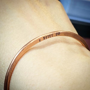 May include: A copper bangle bracelet with the inscription "I STILL DO". The bracelet has a simple, elegant design, suitable for everyday wear. The metal has a warm, reddish-brown hue. The bracelet's diameter is approximately 6.5 cm.