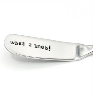 Op de afbeelding: Een roestvrijstalen botermes met de tekst "what a knob!" op het lemmet gestempeld. Het mes heeft een afgeronde punt en een licht gebogen handvat. De tekst is in een eenvoudige, schreefloze lettertype.