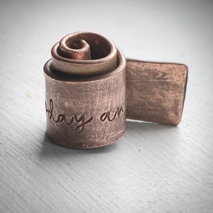 May include: A copper-coloured metal ring with the words "today am" engraved on the cylindrical band. The ring features a spiral design and a rectangular extension. The metal has a textured, brushed appearance.