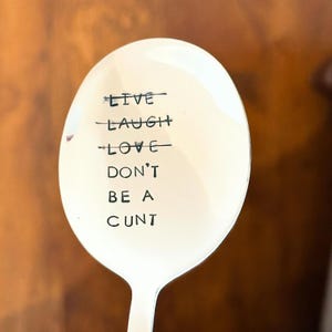 May include: A silver spoon with the words "LIVE LAUGH LOVE DON'T BE A CUNT" etched in black. The spoon is oval-shaped and has a shiny, reflective surface. The text is centered on the spoon's bowl.