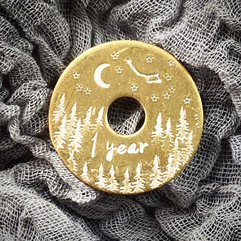 Puede incluir: Un colgante circular dorado con un agujero central. El colgante presenta una escena de bosque con &aacute;rboles blancos, una luna creciente, estrellas y el texto "1 year". El dise&ntilde;o est&aacute; grabado en el metal.
