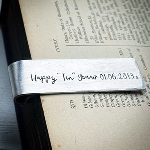 May include: A silver-colored bookmark with the inscription "Happy Tin Years 01.06.2013". The bookmark is curved and rests on the pages of an open book. The text is in a handwritten style.