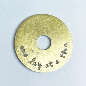 Puede incluir: Un disco met&aacute;lico redondo de color lat&oacute;n con un agujero central. El disco tiene una superficie texturizada y la frase "one day at a time" est&aacute; grabada en cursiva alrededor del borde. Probablemente un art&iacute;culo decorativo o un amuleto.
