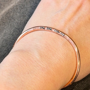 May include: A copper-colored bangle bracelet with etched text. The bracelet is a simple, round band with a slightly textured surface. The etched text appears to be a series of symbols or characters. The bracelet is worn on a wrist.