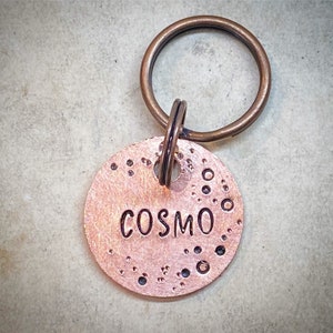 Op de afbeelding: Een koperkleurig huisdierlabel met de naam "COSMO" erop gestempeld. Het label is rond met een reeks kleine, donkere stippen en cirkels langs de randen. Een bronzen sleutelhanger is bevestigd.