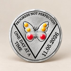 Może przedstawiać: Okrągły srebrny żeton z białą figurą motyla z żółtymi i czerwonymi kropkami pośrodku. Napis PROGRESS NOT PERFECTION, ONE DAY AT A TIME oraz 11.05.2026 jest wygrawerowany czarnymi literami wokół centralnie umieszczonego motywu motyla.