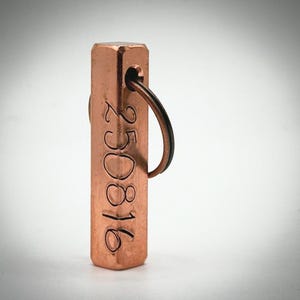 Op de afbeelding: Een rechthoekig koperen label met het gegraveerde nummer "250816" en een zwarte metalen ring. Het label heeft een gat aan de bovenkant voor de ring. Het label heeft een warme koperkleur.