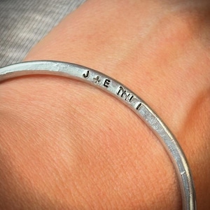May include: A silver bangle with the stamped letters "J + E N I". The bracelet has a hammered texture and is worn on a wrist. The letters are evenly spaced around the bracelet.