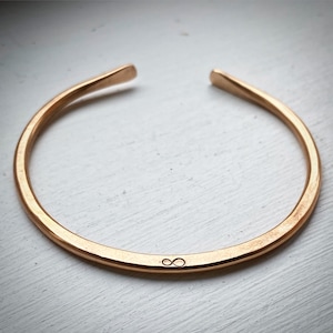 May include: A copper-colored open cuff bracelet. The bracelet has a hammered texture and an engraved infinity symbol. The ends of the bracelet are slightly rounded. The bracelet is shown on a white surface.