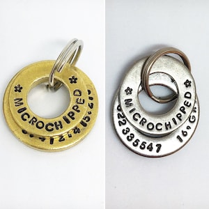 May include: Two metal pet ID tags, one gold-toned and one silver-toned, each with a ring for attachment. The tags are stamped with the words "MICROCHIPPED" and a series of numbers. A paw print design is also stamped on the tags.