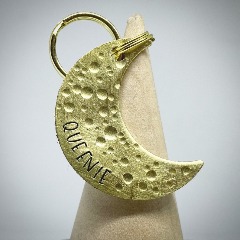 May include: A brass-colored crescent moon-shaped keychain with the name "QUEENIE" stamped on it. The moon has a textured surface with various sized circular indentations. A gold-colored key ring is attached to the top.