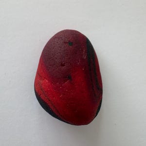 Puede incluir: Una piedra lisa de forma ovalada que presenta un degradado de color rojo vibrante que se transforma en vetas negras profundas. La superficie de la roca muestra texturas naturales y pequeñas hendiduras, con un pigmento rojo saturado.