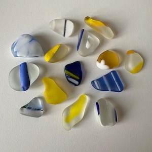 Puede incluir: Una colección de piezas de vidrio de mar de varias formas y tamaños. El vidrio es translúcido con colores como azul, amarillo, blanco y azul marino oscuro. Algunas piezas tienen rayas o bloques de color. El vidrio de mar es liso y redondeado.