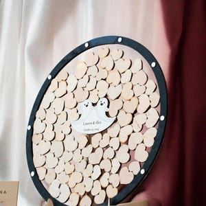 May include: A circular guest book frame with a black border containing numerous small wooden hearts. In the center, a white cutout features two cartoon ghosts and the printed text "Lauren & Alex" followed by the date "October 25, 2024".
