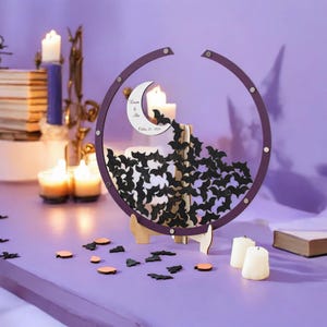 May include: A circular purple wooden frame containing numerous small black bat-shaped cutouts and a white crescent moon cutout featuring the text "Lauren & Alex October 25, 2024". The frame is supported by a light-colored wooden stand on a surface.