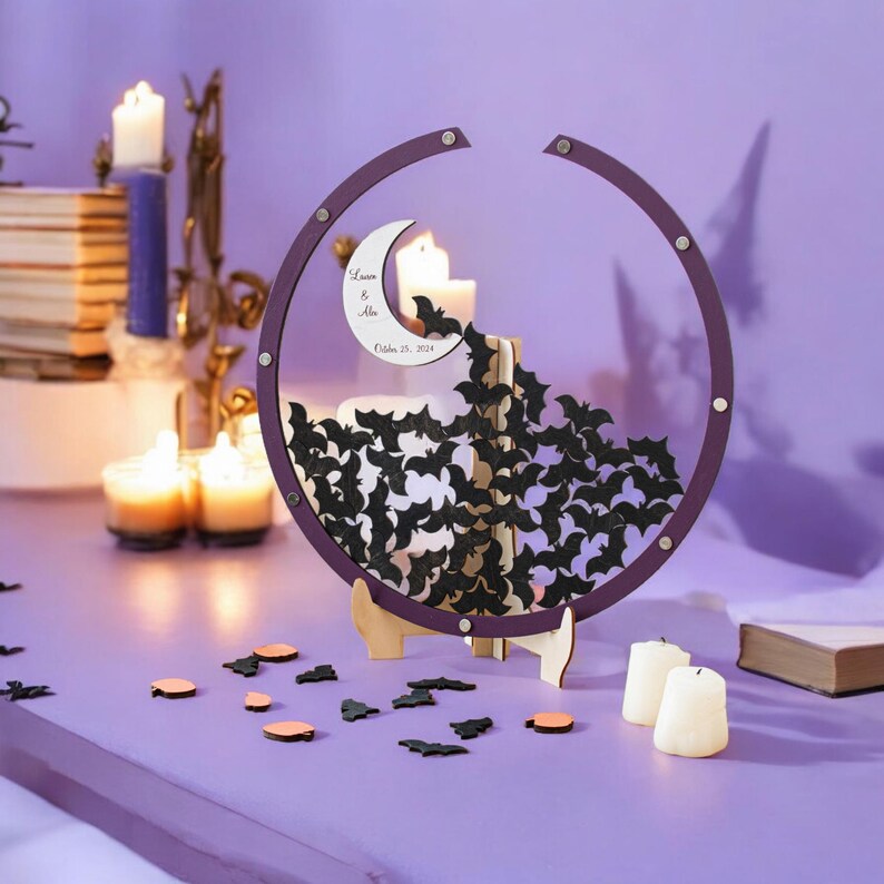 May include: A circular purple wooden frame containing numerous small black bat-shaped cutouts and a white crescent moon cutout featuring the text "Lauren & Alex October 25, 2024". The frame is supported by a light-colored wooden stand on a surface.