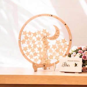 May include: A wooden wedding guest book alternative with a clear circular frame, a crescent moon, and star-shaped pieces. The frame holds numerous wooden stars. A small sign reads "Please Sign a Star."
