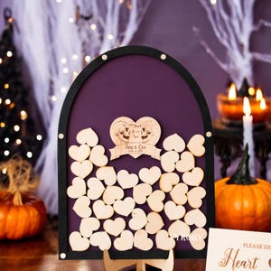 Könnte beinhalten: Ein violettes und schwarzes Gästebuch aus Holz mit einem Totenkopf- und Knochenkreuz-Design. Der Text "Till Death Do Us Part" ist in das herzförmige Schild graviert. Das Schild enthält auch die Namen "Doug & Chris" und das Datum "October 11, 2018".