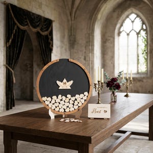 May include: A circular wooden guest book frame with a black interior, filled with small wooden hearts. A central wooden emblem features wings and text. A small rectangular sign reads "Please sign a Heart" with a heart icon next to it.