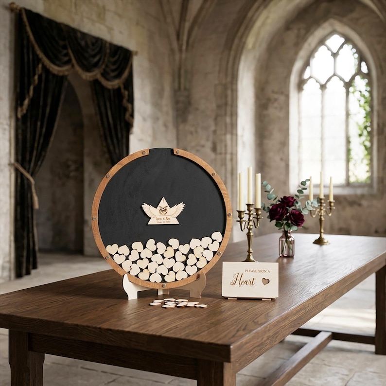 May include: A circular wooden guest book frame with a black interior, filled with small wooden hearts. A central wooden emblem features wings and text. A small rectangular sign reads "Please sign a Heart" with a heart icon next to it.