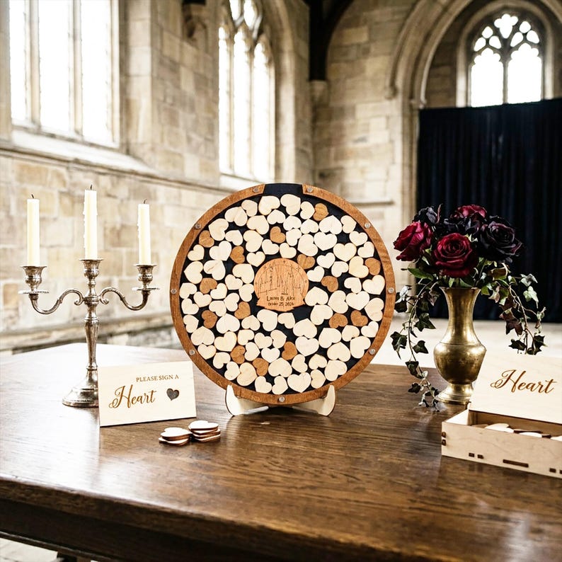 May include: A circular wooden guest book frame filled with small wooden hearts, featuring a central engraved plaque with the text "Laura & Alex October 25, 2024". Includes a silver candelabra, a sign reading "Please Sign A Heart", and a wooden box.
