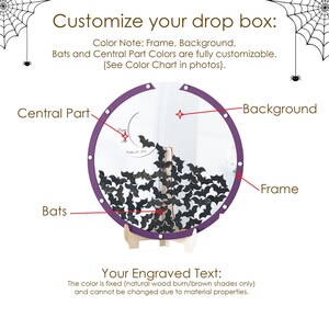 May include: A circular drop box with a purple frame, a clear background, and numerous black bat-shaped cutouts inside. The display features a central part with engraved text, supported by a light-colored wooden stand. Spider web graphics appear in the top corners.