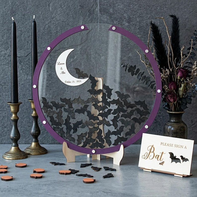 May include: A circular clear acrylic guest book frame with a purple border, featuring a white crescent moon with the text "Lauren & Alex October 25, 2024" and numerous black wooden bat-shaped tokens inside, supported by a light wood stand.