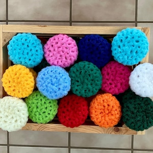May include: A wooden crate filled with a variety of colorful, crocheted scrubbies. The scrubbies are in shades of blue, pink, yellow, green, red, orange, and white. The scrubbies are round and have a textured appearance.