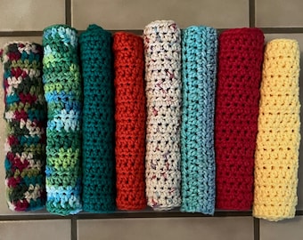 May include: Eight cylindrical, hand-crocheted cloths arranged in a row. The colors include a multi-colored variegated yarn, a blue and green mix, solid teal, solid red, a cream speckled yarn, solid light blue, solid dark red, and solid yellow.