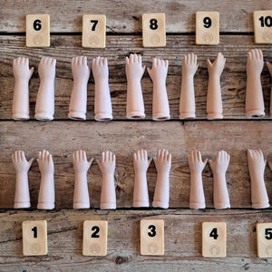 May include: A collection of antique doll hands and numbered tiles arranged on a wooden surface. The hands are pale pink and in various poses. The tiles are cream-colored with black numbers from 1 to 10.