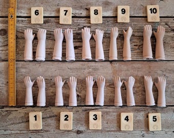 May include: A collection of antique doll hands and numbered tiles arranged on a wooden surface. The hands are pale pink and in various poses. The tiles are cream-colored with black numbers from 1 to 10.