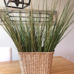 May include: A tall, green, artificial grass plant in a woven wicker basket. The basket is light brown and has a rectangular shape.