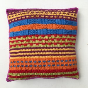 May include: A colorful knitted pillow with a geometric pattern in shades of orange, yellow, red, green, blue, and purple. The pillow has a pink border.